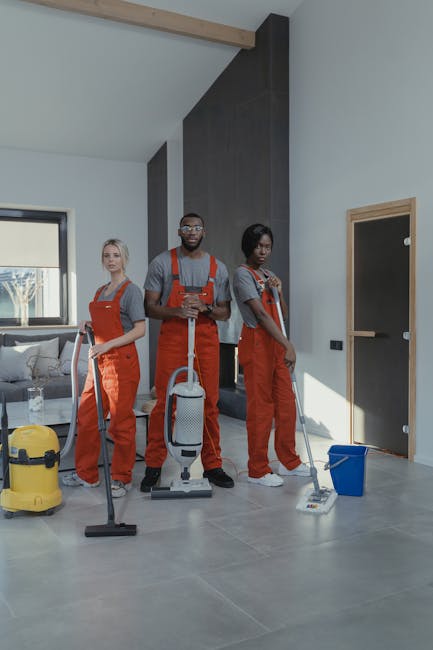Three professional cleaners, wearing grey shirts and bright orange overalls, are standing in a modern, well-lit living room with polished grey tiled floors. They are holding cleaning equipment, includ