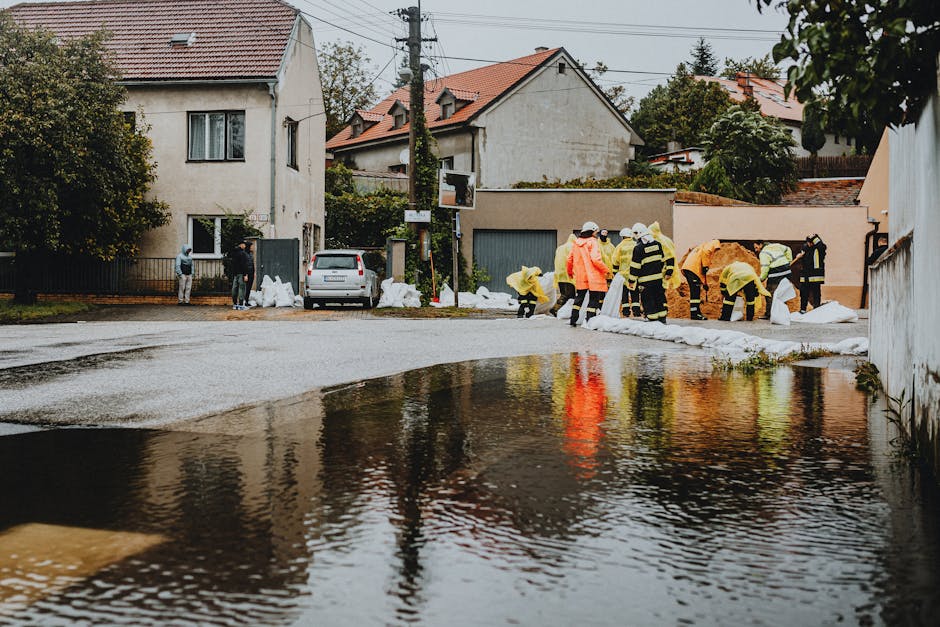 A red firefighting truck labeled 'BOMBEROS' is partially submerged in floodwater outside a residential area. The truck is equipped with extensions for water spraying, with the surrounding street and n