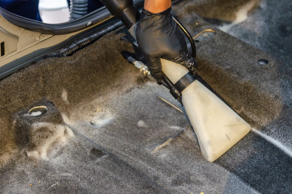 A person using a handheld electric upholstery cleaner with a yellow and black body is deep cleaning a dark grey fabric sofa in a domestic living room. The individual is wearing black gloves and black